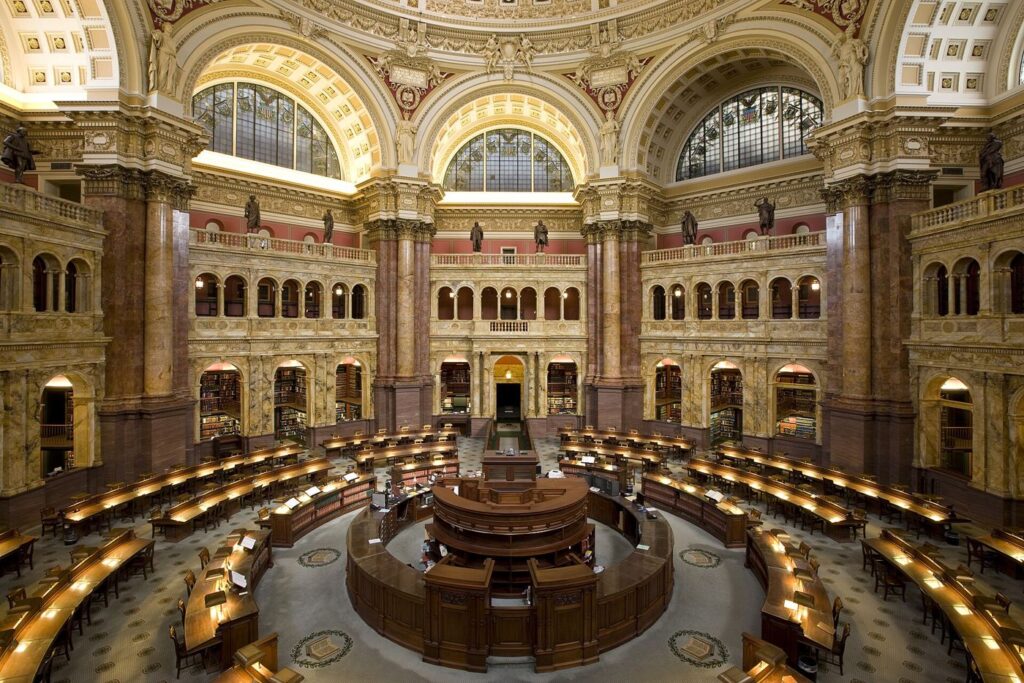 Readers study quietly at long wooden tables inside a bright public library reading room.